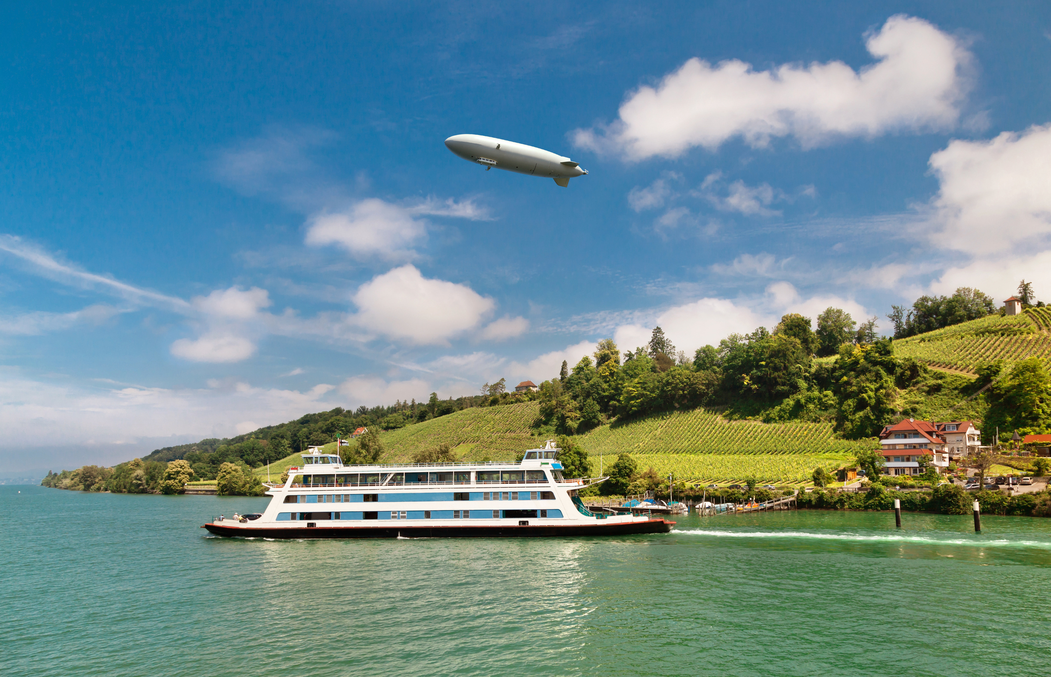 A Zeppelin hovers over a river landscape with vineyards and a ferry.