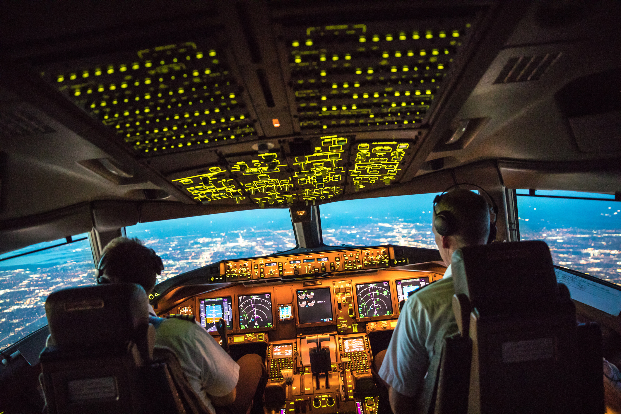 An illuminated aircraft cockpit with two pilots approaching a city in the evening.