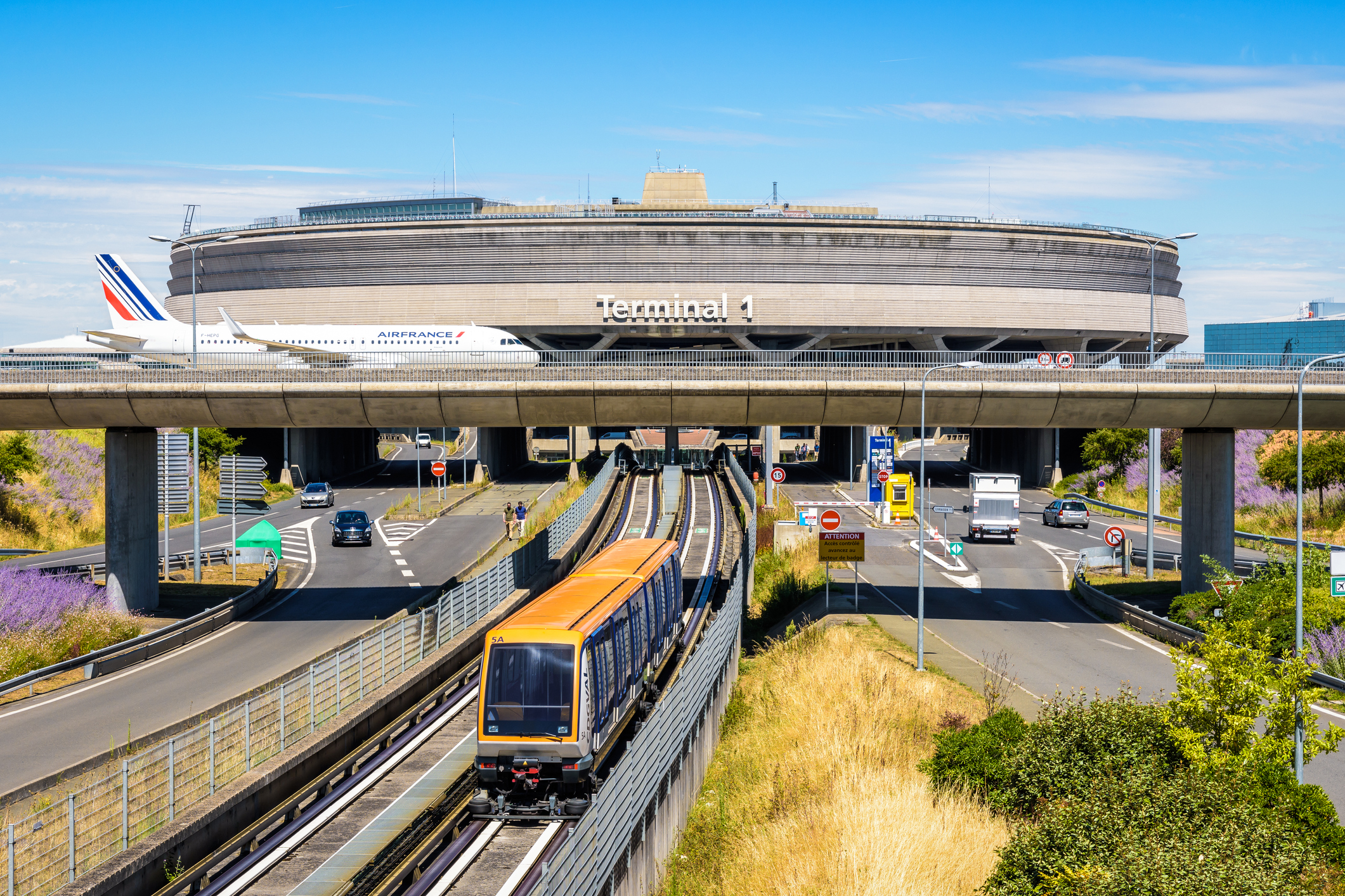 An image of Terminal 1 of Charles de Gaulle Airport, a streetcar can be seen in the foreground, an Air France plane is crossing a highway on a bridge in the background.