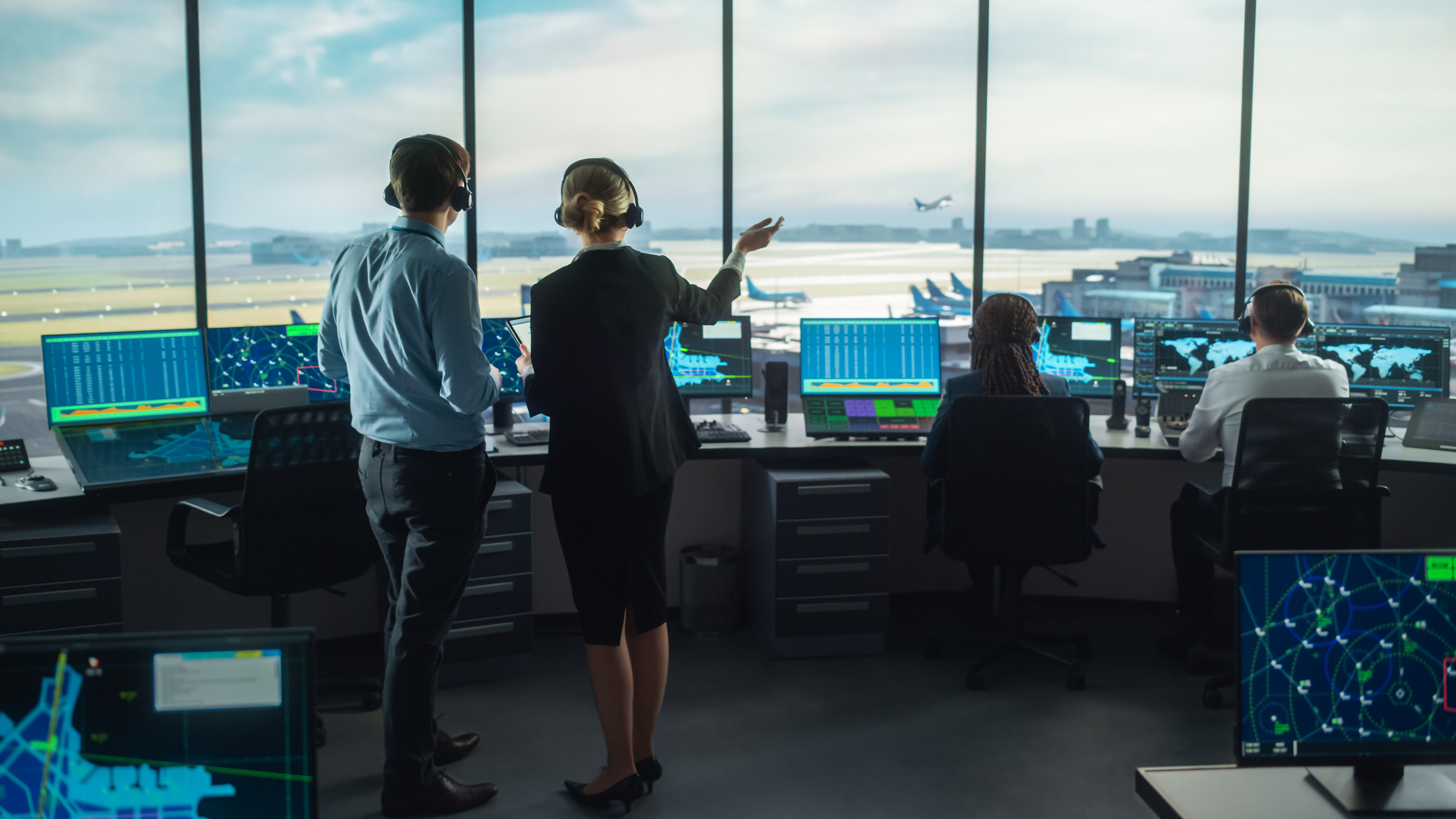 Inside an airport tower there are several air traffic controllers and a few screens; in the background, runways and a plane taking off can be seen through the window front.