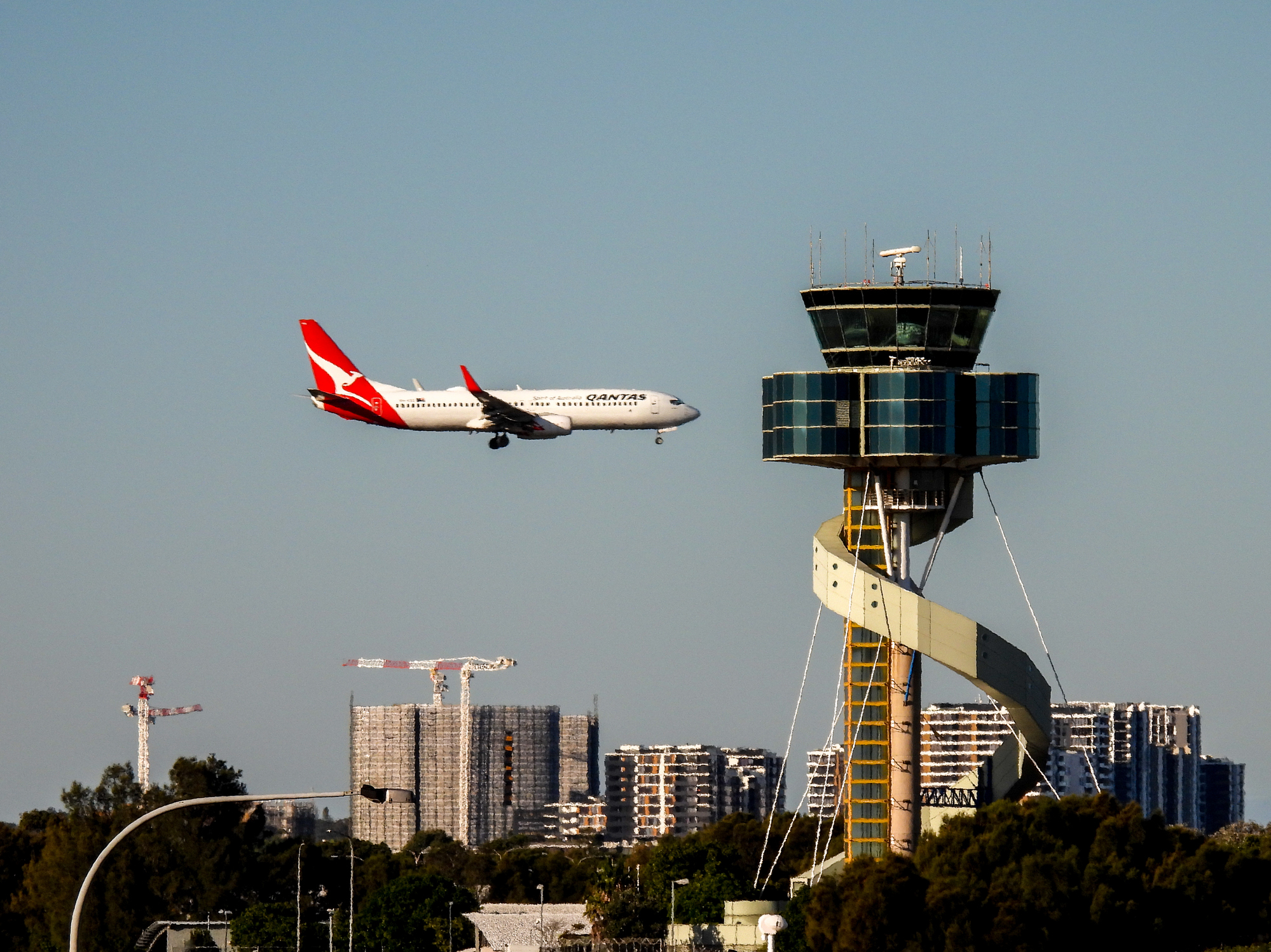 A Qantas plane is approaching Sydney Airport, with the striking airport tower further back.