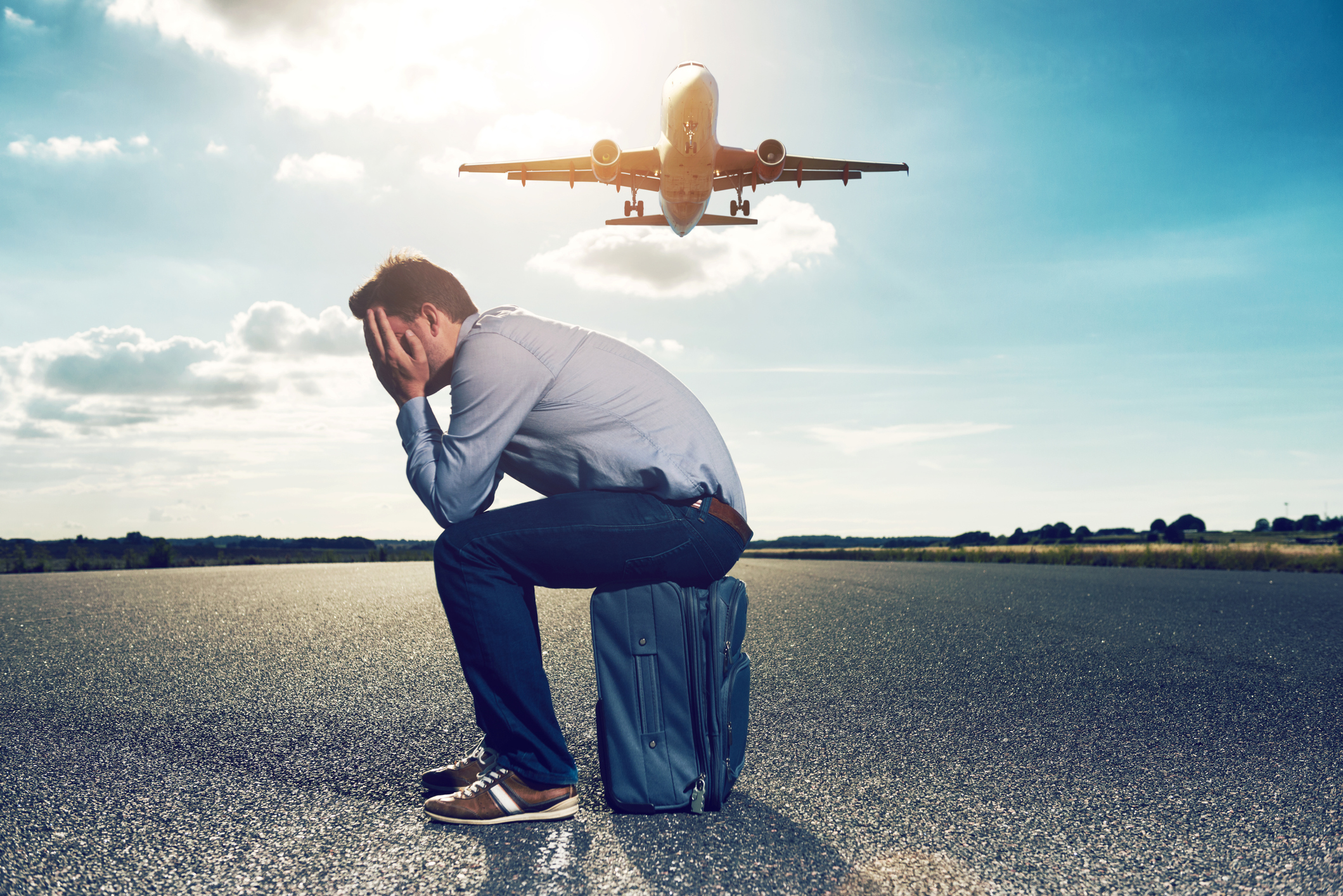 A man is sitting on a blue suitcase with his hands in front of his face while an aeroplane flies behind him.