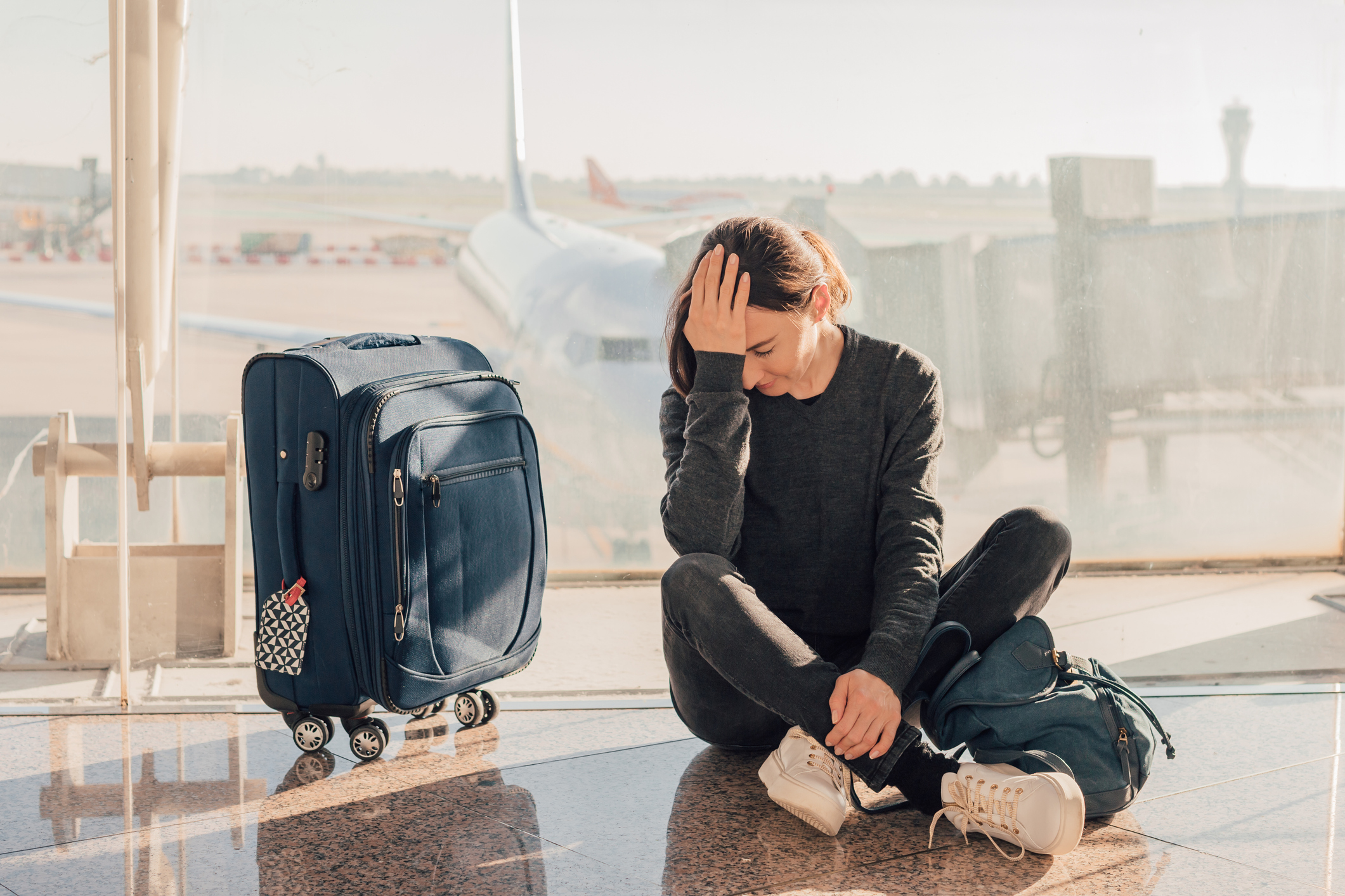 A woman sitting in the airport next to her suitcase and with her backpack on the floor puts one hand on her forehead.