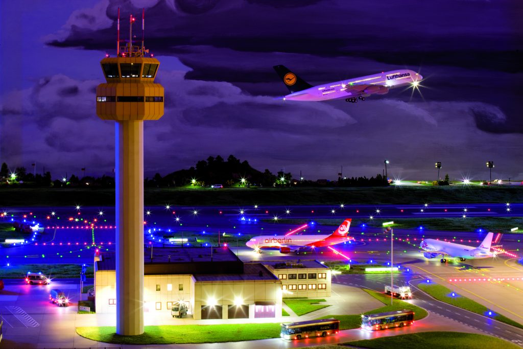The tower of Knuffingen Airport at night, with a Lufthansa plane taking off in the background and two other planes heading to the tarmac.