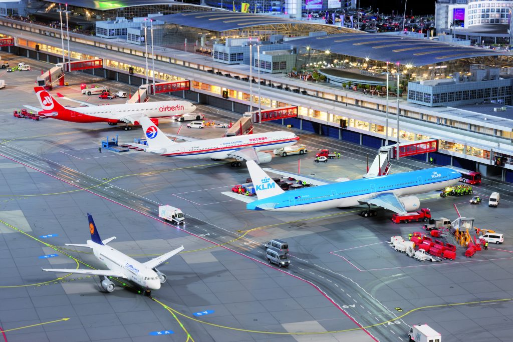 Several airplanes are parked with docked gangways at the gates, in the foreground a Lufthansa plane is taking off, in the background are the terminals.