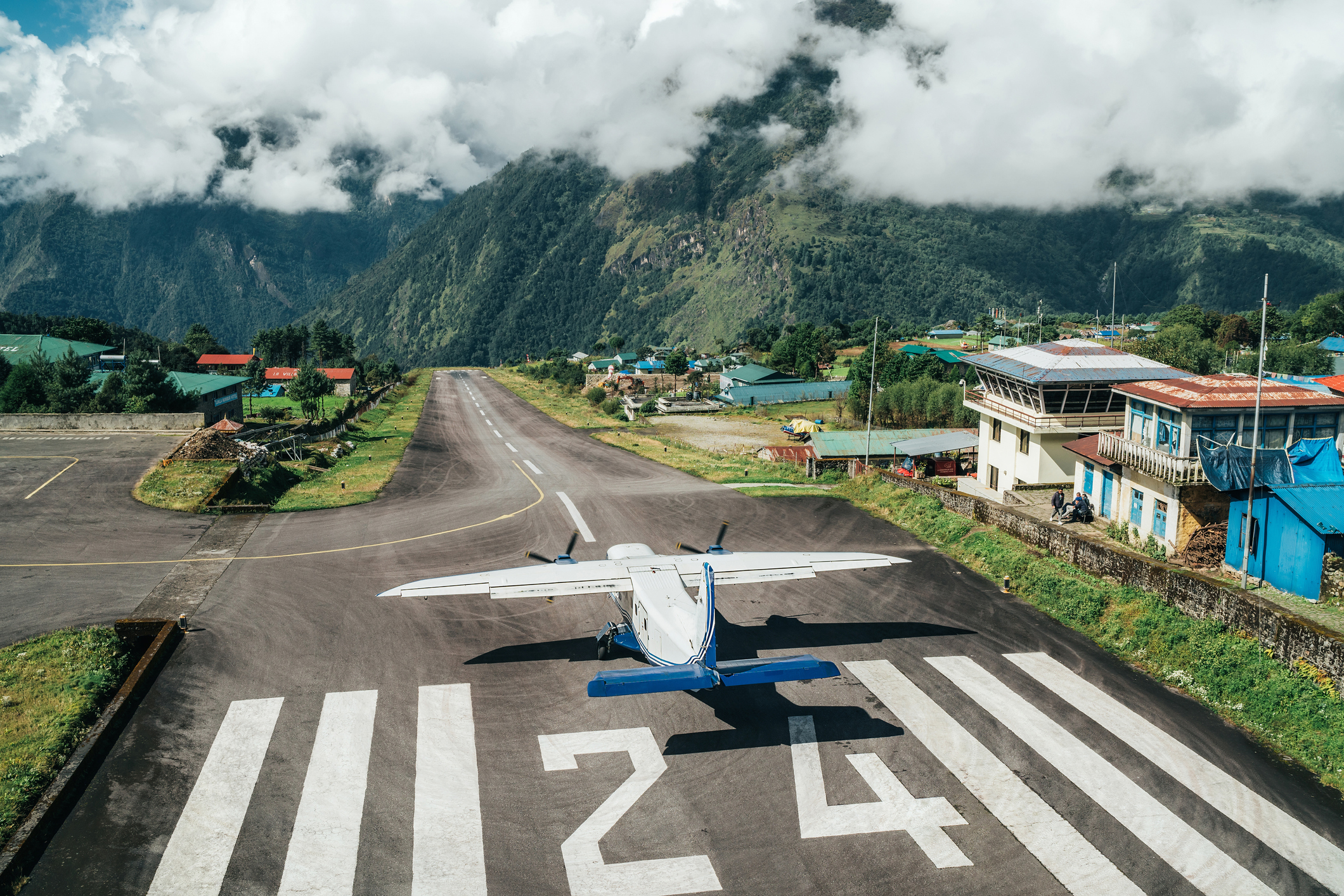 The runway in Lukla with sunshine but also some low-hanging clouds, a propeller plane is just starting its takeoff.