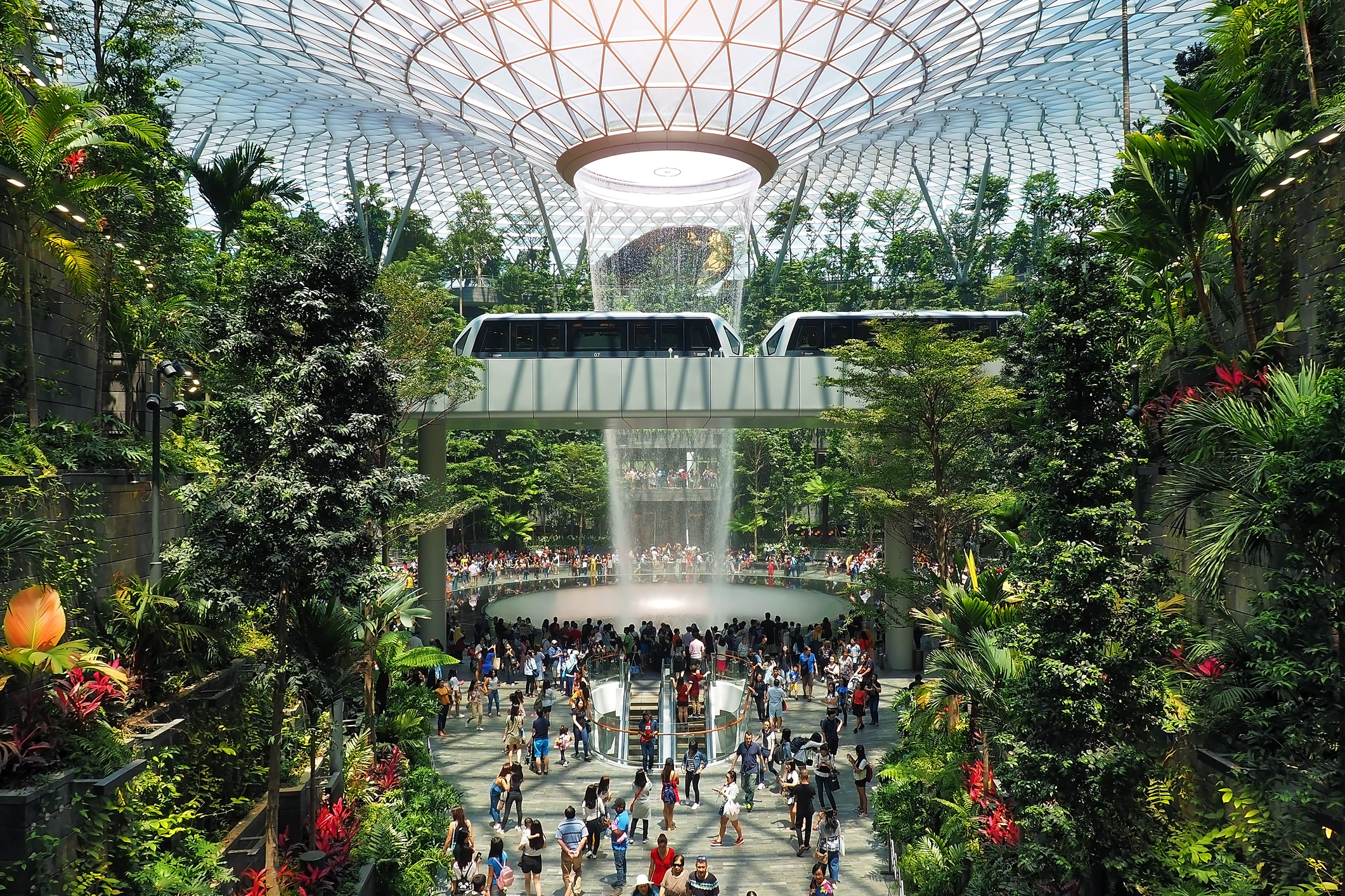 A panoramic shot from inside the Jewel Changi, showing the Rain Vortex waterfall, tropical plants and many travellers.