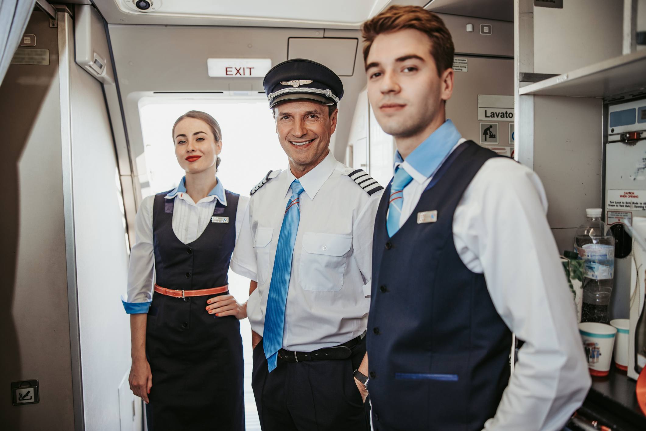 A pilot and two service personnel stand in front of the exit of an airplane and smile into the camera.