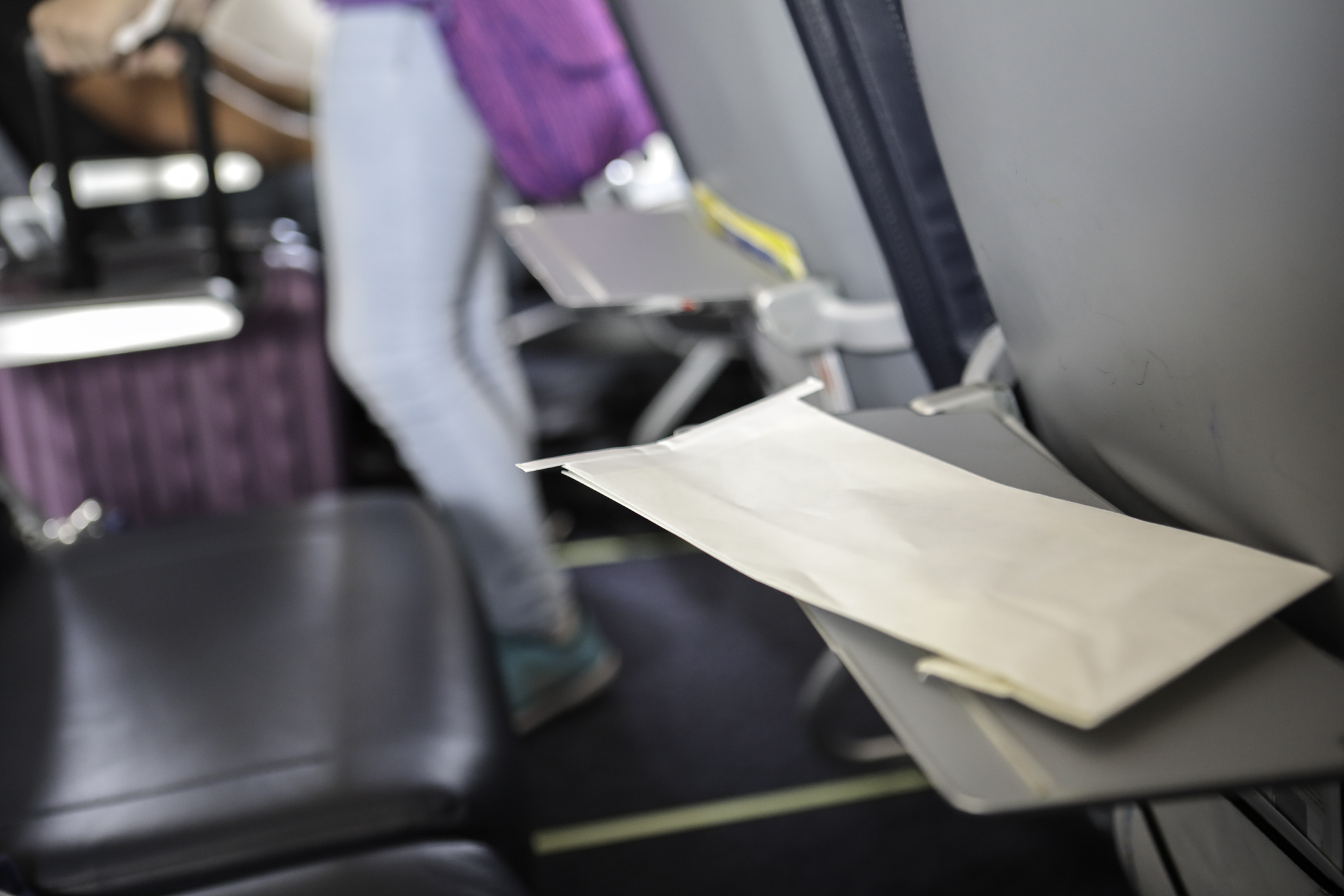 An envelope is lying on a folded down table at an airplane seat, in the background there is another folded down table and a person on the aisle with suitcase.