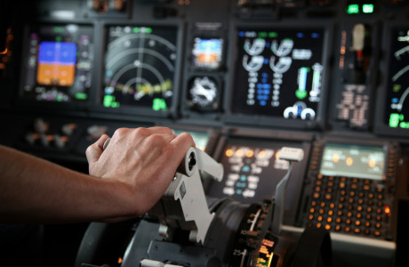 A hand pushes a control forward, with numerous gauges and meters of a cockpit in the background.