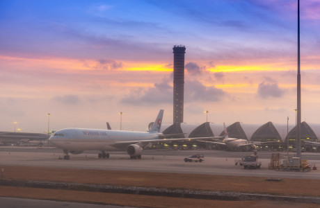 At dusk, a Korean Air plane stands on the tarmac in the foreground, while the airport tower at Bangkok Suvarnabhumi Airport rises into the air in the background.